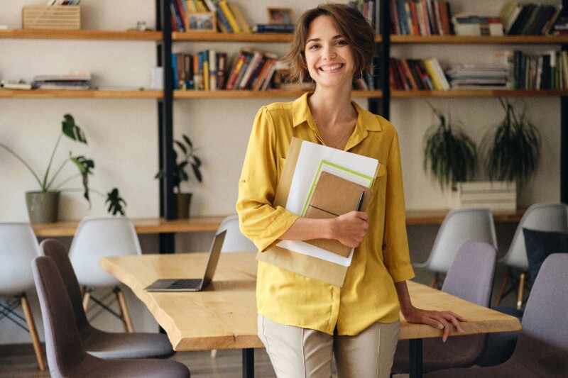 Young cheerful woman in yellow shirt leaning on desk with notepad and papers in hand joyfully looking in camera in modern office
