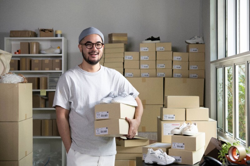 Confident young Asian man retail seller, entrepreneur, online store drop shipping small business owner looking at camera standing in delivery shipping warehouse with parcel boxes.
